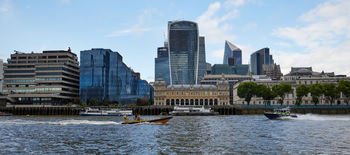 This urban landscape photograph depicts a police boat navigating the River Thames in London during a summer afternoon. The main subject, a police boat, is positioned on the river with the iconic Walkie Talkie building—also known as the Sky Garden—clearly visible in the background along with other notable examples of modern and historic London architecture. The riverside scene showcases a mix of glass-fronted skyscrapers and traditional classical buildings, highlighting the unique blend of old and new that characterizes London’s urban environment along the Thames. The river is animated with other vessels, and the waterfront is lined with trees, further adding to the lively summer atmosphere in this part of the city.